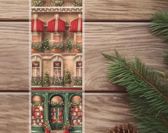 May include: A Christmas-themed bookmark featuring a detailed illustration of a building facade. The building has red awnings, decorated windows, and a bookstore on the ground floor. The bookmark is set against a wooden background with pine needles and a pine cone.