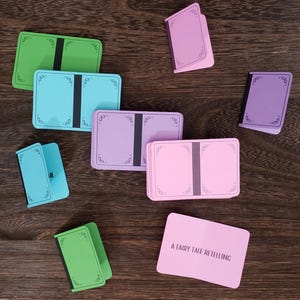 May include: A set of blank cards with decorative borders, featuring a variety of colors, including green, blue, pink, and purple. The cards are arranged on a wooden surface. The text "A Fairy Tale Retelling" is printed on a pink card.