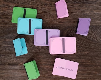 May include: A set of blank cards with decorative borders, featuring a variety of colors, including green, blue, pink, and purple. The cards are arranged on a wooden surface. The text "A Fairy Tale Retelling" is printed on a pink card.
