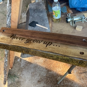 May include: A wooden shelf with the engraved phrase "Never grow up..." is shown. The shelf has two pre-drilled holes and a natural edge. The wood has a two-tone finish, with a darker stain on top and a lighter, patterned edge.