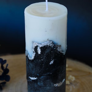 Puede incluir: Una vela cilíndrica con la parte superior blanca y una base azul oscuro y negra. Acentos de papel de plata acentúan la transición entre los colores. La vela tiene una mecha blanca y mide unos 10 cm de altura.