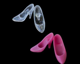 May include: A pair of clear high-heeled shoes with embedded iridescent glitter and a pair of solid pink high-heeled shoes. Each shoe features a pointed toe, a curved heel, and a smooth, molded construction, presented as four individual items.