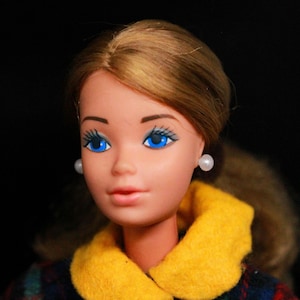 Puede incluir: Una muñeca vintage con cabello castaño, ojos azules y un cuello amarillo. Lleva un vestido a cuadros y pendientes de perlas.