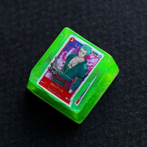 May include: A green, translucent, square-shaped keycap with a printed image of a cartoon character with green hair and a red sash. The text "5000" and "3" are printed on the keycap. The keycap is made of resin and has a glittery finish.