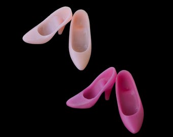 May include: Two pairs of miniature high-heeled shoes, one pair in a light pastel pink and the other in a vibrant magenta. Each shoe features a pointed toe, a smooth molded finish, and a small heel, presented against a solid black background.