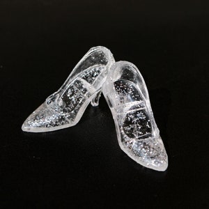 May include: Two clear glass slipper shoes with silver glitter. The shoes have a pointed toe and a high heel.