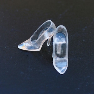 May include: Two clear, miniature high-heeled shoes. The shoes are made of transparent material, with pointed toes and slender heels. The heels are approximately 1 inch tall. The shoes are positioned on a dark surface.