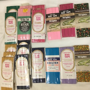May include: Assortment of single fold bias tape in various colors, including green, pink, purple, blue, and gold. The tape is labeled with brand names like Coats, Wrights, and Boiltex. Each spool contains 4 yards of bias tape.