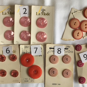 May include: Assortment of vintage buttons in various colors and styles, including pink, red, and brown. The buttons are arranged on cards with numbers and text, such as "La Mode" and "Streamline".