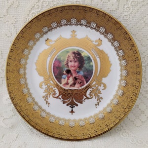 Może przedstawiać: Biały talerz porcelanowy z pozłacanym rantem i misternymi złotymi wzorami. W centrum znajduje się portret młodej dziewczyny trzymającej szczeniaka, otoczony złotym wzorem kwiatowym.