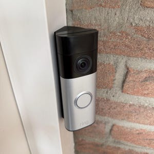 Op de afbeelding: Een rechthoekige videodeurbel met een zwart bovengedeelte met een cameralens en een zilverkleurig ondergedeelte met een ronde knop en het woord ring in kleine letters. Het apparaat is verticaal gemonteerd op een wit deurkozijn.