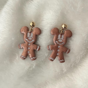 May include: A pair of earrings featuring a gingerbread man design with a Mickey Mouse silhouette. The earrings are a light brown color with white accents and gold-colored posts.