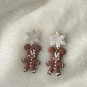 May include: A pair of brown gingerbread man earrings with white icing details, topped with white snowflake charms. The earrings are designed with a festive, holiday theme, perfect for seasonal wear.