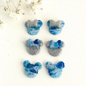 May include: Six blue and silver mouse-shaped earrings with glitter accents. The earrings have a marbled blue design and are arranged on a white surface. Some earrings feature a bow.