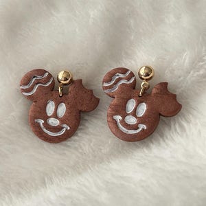 May include: A pair of brown Mickey Mouse-shaped earrings with white accents. The earrings have a gold-colored ball at the top and a smiling face design. The earrings are on a white, fluffy surface.
