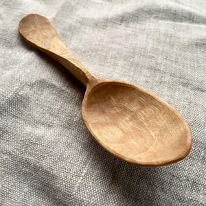 Könnte beinhalten: Ein Holzlöffel mit hellbrauner Farbe und einer glatten, abgerundeten Oberfläche. Der Löffel ist aus Naturholz gefertigt und hat ein klassisches Design.