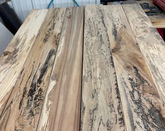 May include: A collection of unfinished wooden planks with unique grain patterns. The wood features a mix of light tan, cream, and dark brown hues, with striking black streaks and knots. The planks are arranged side by side, showcasing the natural wood texture.