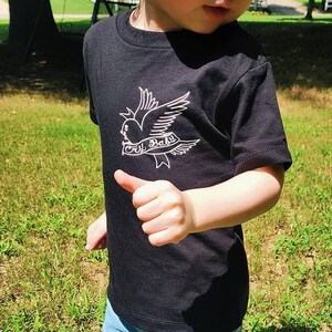 Op de afbeelding: Een peuter draagt een zwart T-shirt met een witte duif en de tekst "Lil Baby" erop. De peuter draagt ook een blauwe spijkerbroek, kleurrijke sokken en zwart-wit geblokte instappers.