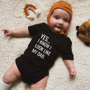 May include: A baby wearing a black bodysuit with the text "YES... I KNOW I LOOK LIKE MY DAD." The baby is wearing a brown hat and is lying on a white fluffy surface.