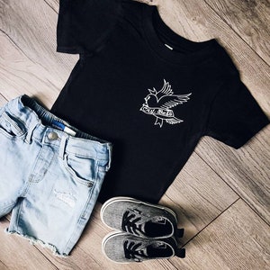 Op de afbeelding: Zwart T-shirt met een witte afbeelding van een vogel met een hart en de tekst "cry baby". Het shirt wordt gecombineerd met een blauwe denim short en grijze sneakers.