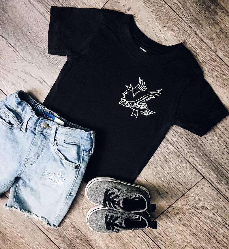 Op de afbeelding: Zwart T-shirt met een witte afbeelding van een vogel met een hart en de tekst "cry baby". Het shirt wordt gecombineerd met een blauwe denim short en grijze sneakers.