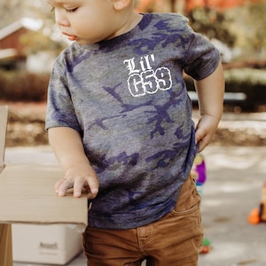 May include: A young child wearing a blue and green camouflage t-shirt with the text "Lil' 653" in white. The child is wearing brown corduroy pants.