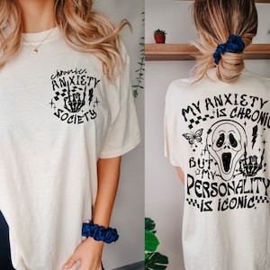 May include: Cream-colored t-shirt with black graphic text and illustrations. The front reads "Chronic Anxiety Society" with a skeleton hand. The back features a Scream mask and the text "My Anxiety is Chronic But My Personality is Iconic."