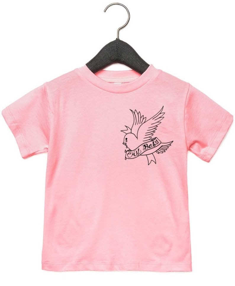 Op de afbeelding: Roze T-shirt met korte mouwen met een zwart-witte afbeelding van een vogel die een hart vasthoudt met de tekst "cry baby" erop.