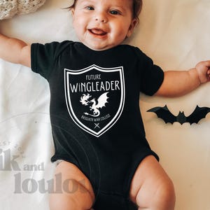 May include: A baby wearing a black bodysuit with a white graphic that says "Future Wingleader" and a dragon illustration. The graphic is inside a shield shape with the text "Bassgath War College" underneath the dragon.