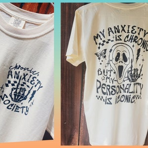 Może przedstawiać: Kremowa koszulka z czarnym tekstem graficznym i ilustracjami. Prz&oacute;d przedstawia "Chronic Anxiety Society" z gestem dłoni. Tył m&oacute;wi "My Anxiety is Chronic But My Personality is Iconic" z maską Scream.