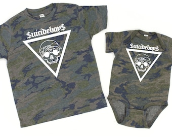 Puede incluir: Dos camisetas a juego con estampado de camuflaje. Una es una camiseta de manga corta y la otra es un body para bebé. Ambas tienen un triángulo blanco con un gráfico de calavera y el texto "$uicideboy$".