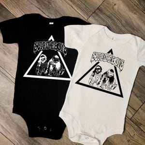 May include: Two black and white t-shirts with a triangle graphic and the text "Suicideboys". The shirts are on a wooden surface.