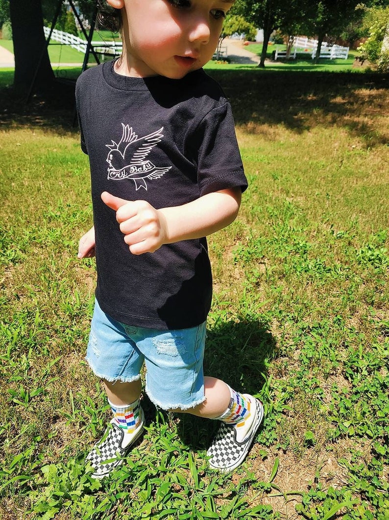 Op de afbeelding: Een peuter draagt een zwart T-shirt met een witte duif en de tekst "Lil Baby" erop. De peuter draagt ook een blauwe spijkerbroek, kleurrijke sokken en zwart-wit geblokte instappers.