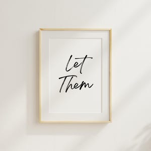 以下が含まれることがあります： 白い背景に黒い筆記体で「Let Them」と書かれた額装されたプリント。フレームは明るい木の色です。