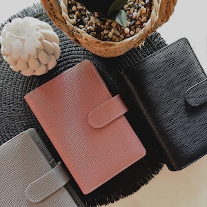 May include: Three rectangular planners with a textured, horizontal ribbed surface. Each planner features a rounded tab closure on the right side. The planners are coloured in muted grey, dusty rose, and solid black, arranged in a diagonal cluster.