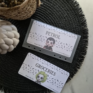 May include: Two white budget cards with cartoon character illustrations and the words "PETROL" and "GROCERIES" printed on them. The cards are decorated with bat silhouettes. One card is in a clear plastic sleeve. The cards are on a black textured surface.
