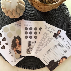 May include: A selection of savings challenge cards with different designs and text, including "Make It Happen Savings" and "The $2 Coin Challenge." The cards feature illustrations and a decorative candle.