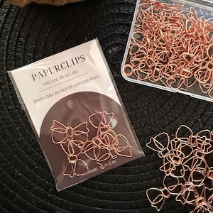 May include: A collection of rose gold metal paperclips shaped like bows. Some clips are contained within a clear plastic storage box, while others are inside a transparent retail bag labelled PAPERCLIPS DREAM. PLAN. DO. BOSS GIRL BUDGETS ACCESSORIES.