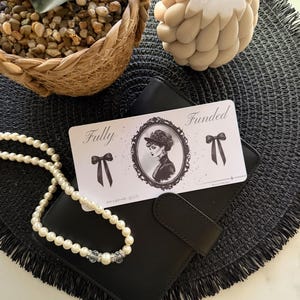 May include: A black wallet with a pearl necklace and a card that reads "Fully Funded" with a vintage illustration. A decorative candle and a woven basket with small stones are also in the image. The items are arranged on a black textured mat.