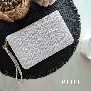 May include: A white, textured clutch bag with a wrist strap and gold-coloured hardware. The clutch has a rectangular shape and a ridged pattern. The text "#LILI" is visible. The clutch is on a black, fringed mat.