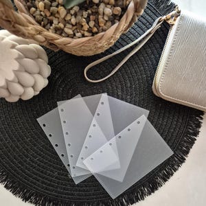 May include: A collection of clear, square, plastic binder pockets with reinforced holes, arranged on a black, textured surface. A woven basket with pebbles, a white decorative object, and a white wallet with a wrist strap are also visible.