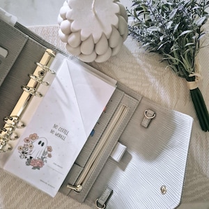 May include: A light grey textured personal planner with a gold six-ring binder mechanism. Inside is a white paper insert featuring a ghost illustration surrounded by flowers and the text "NO COFFEE NO WORKEE" printed in black ink.