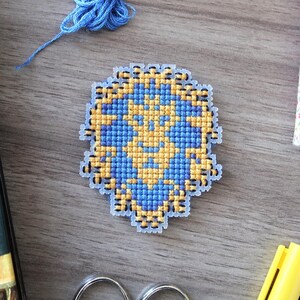 May include: A cross-stitch emblem in blue and yellow, featuring a detailed design. The emblem is surrounded by craft supplies, including blue and yellow thread, scissors, and markers. A book with a blue cover and a floral-patterned notebook are also present.