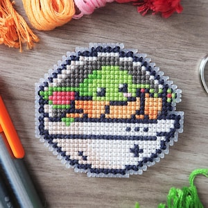 May include: A cross-stitch design of a green character in a white and blue pod. The image includes orange, pink, and green embroidery threads, pink and orange markers, and silver scissors. The craft supplies are arranged on a wooden surface.