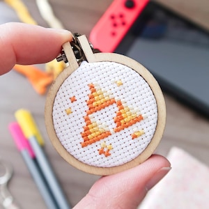 May include: A small, circular cross-stitch embroidery hoop featuring a geometric design in orange and yellow. The wooden hoop is held in a hand, with a Nintendo Switch console in the background. Craft supplies are also visible.