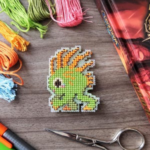 May include: A cross-stitch design of a green creature with orange hair, next to a book with a gold title. Colorful embroidery floss in orange, blue, green, and pink, along with scissors and markers, are also visible.