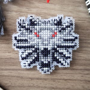 May include: A cross-stitch design of a wolf's head with red eyes, in shades of gray and black. The design is on a light-colored plastic canvas. Yarn tassels and a book are in the background.