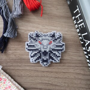 May include: A cross-stitched emblem featuring a wolf's head with red eyes, in shades of gray and black. The emblem is on a wooden surface, with strands of thread in gray and red, and a book titled "The Last Wish".