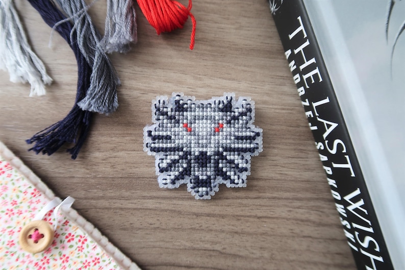 May include: A cross-stitched emblem featuring a wolf's head with red eyes, in shades of gray and black. The emblem is on a wooden surface, with strands of thread in gray and red, and a book titled "The Last Wish".