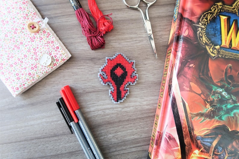 May include: A collection of craft supplies and a book are arranged on a wooden surface. Items include a cross-stitched emblem in red, black, and gray, embroidery floss, scissors, pens, and a floral notebook with a button closure.